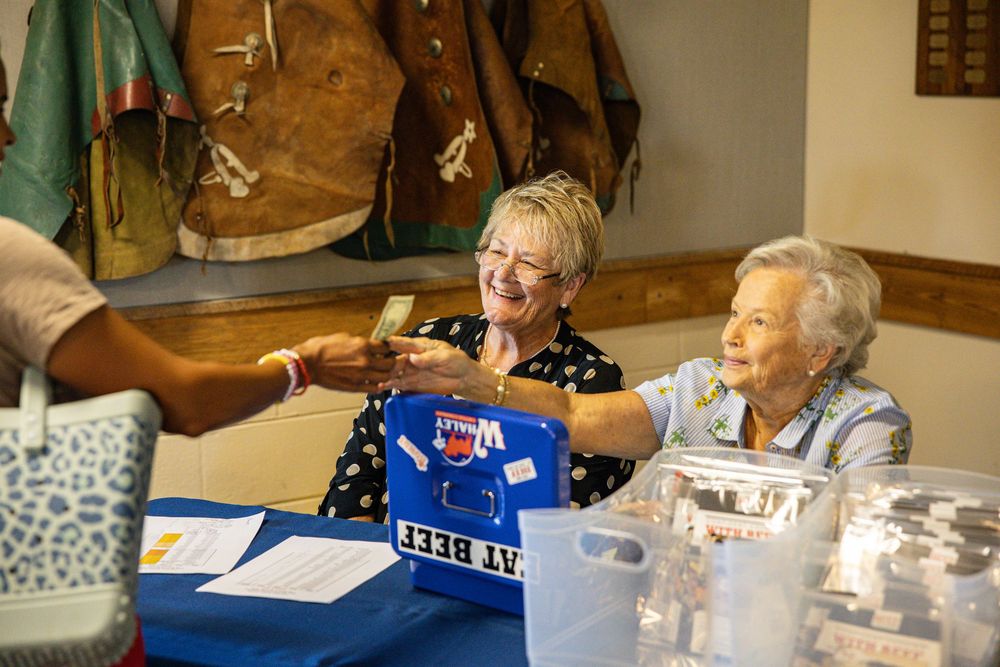 Two elderly women smile while handing items across a registration table at an indoor community event.