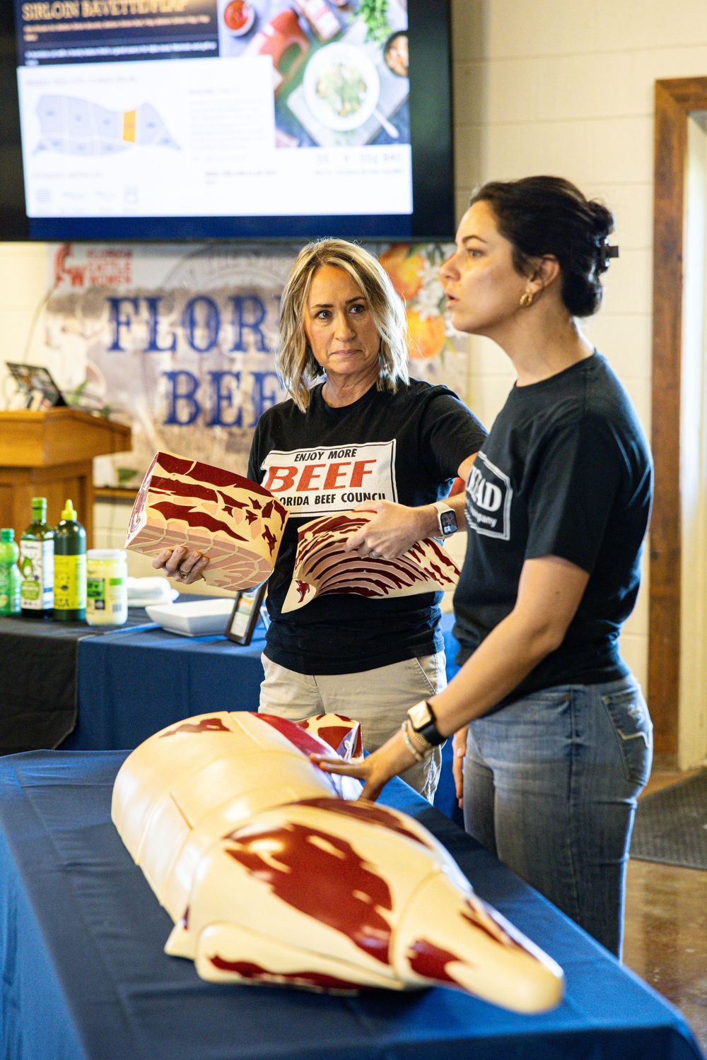 Two women in Florida Beef Council shirts present a beef cut model at an educational display event.