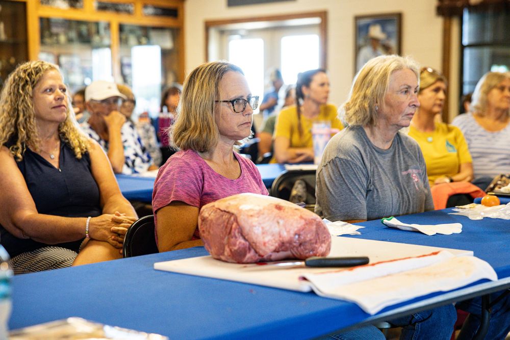 Women attending a cooking class with a large cut of raw meat on a cutting board at a blue table.
