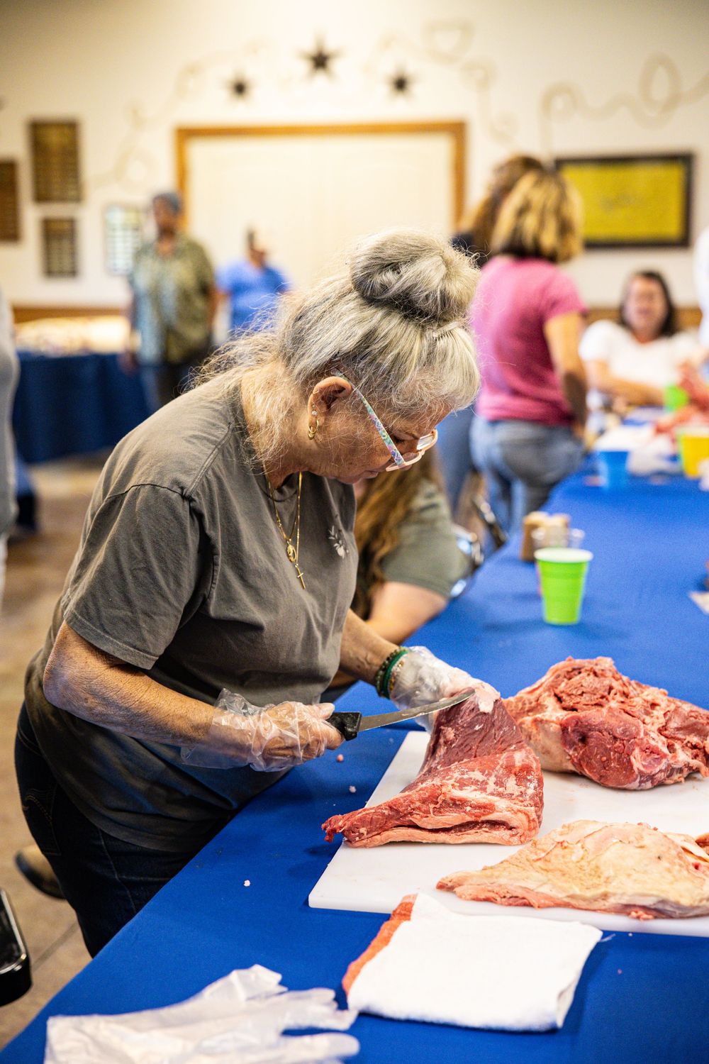Elderly woman with gray hair cutting raw meat on a white cutting board at a blue table community event.
