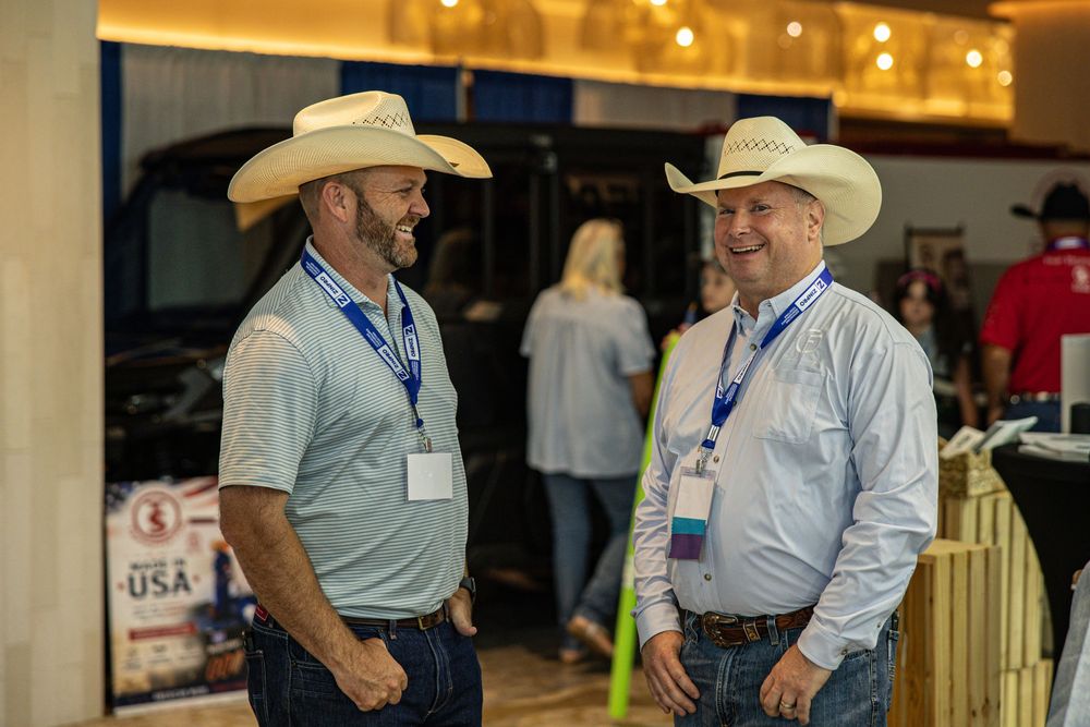 Two men in cowboy hats and lanyards smile and chat at an indoor USA-themed networking event.