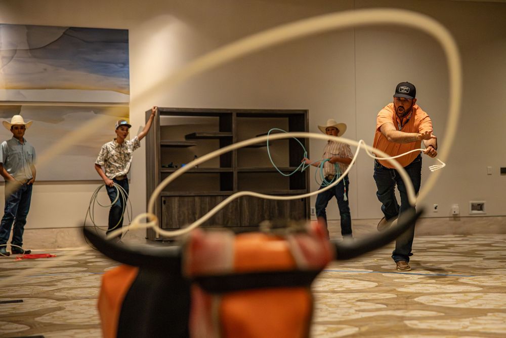 Cowboys practice roping skills indoors, targeting a longhorn dummy in a conference room setting.