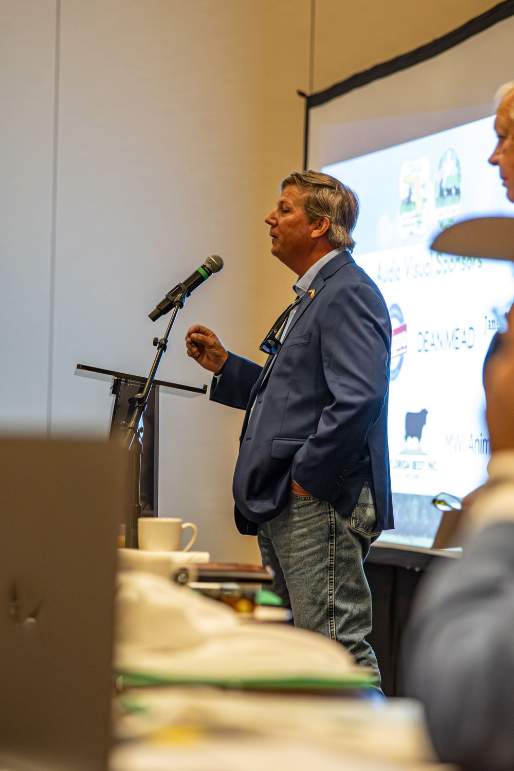 Man in blue blazer speaking at podium with microphone during a business conference presentation.