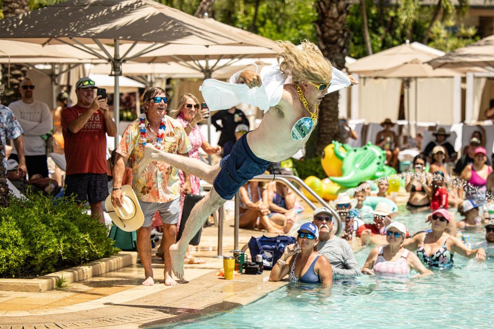 Person doing a backflip into a crowded resort pool surrounded by spectators in tropical attire on a sunny day.