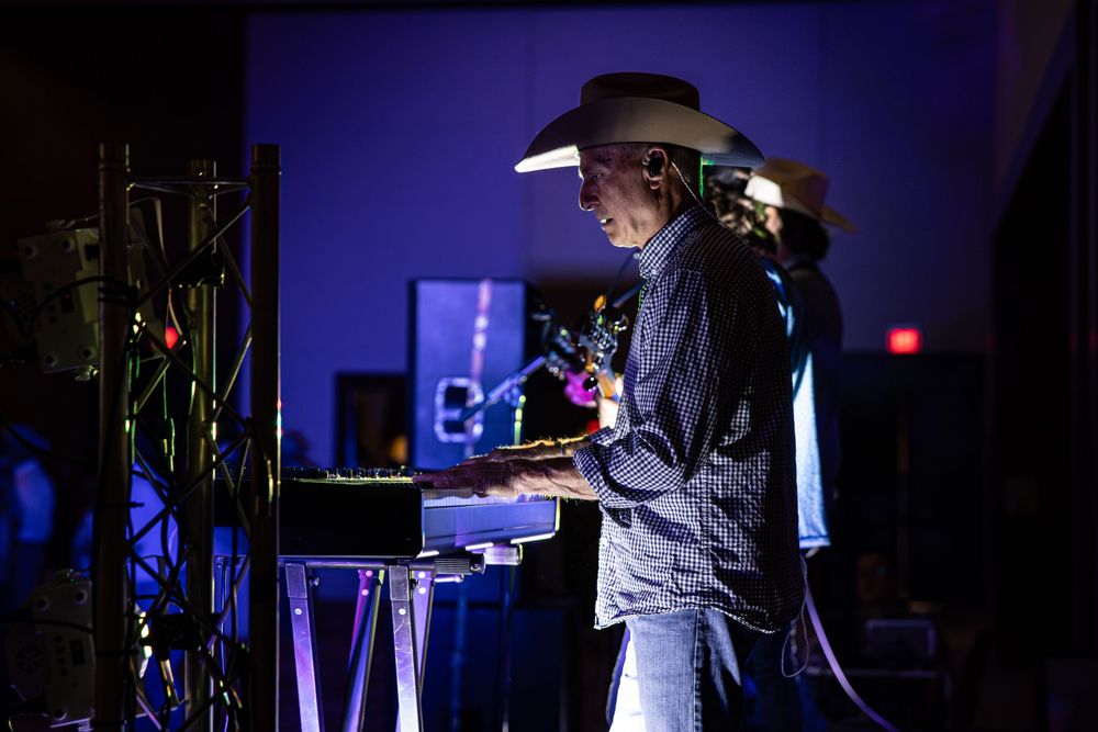 A DJ wearing a cowboy hat performs at a dimly lit event, illuminated by blue and purple stage lighting.