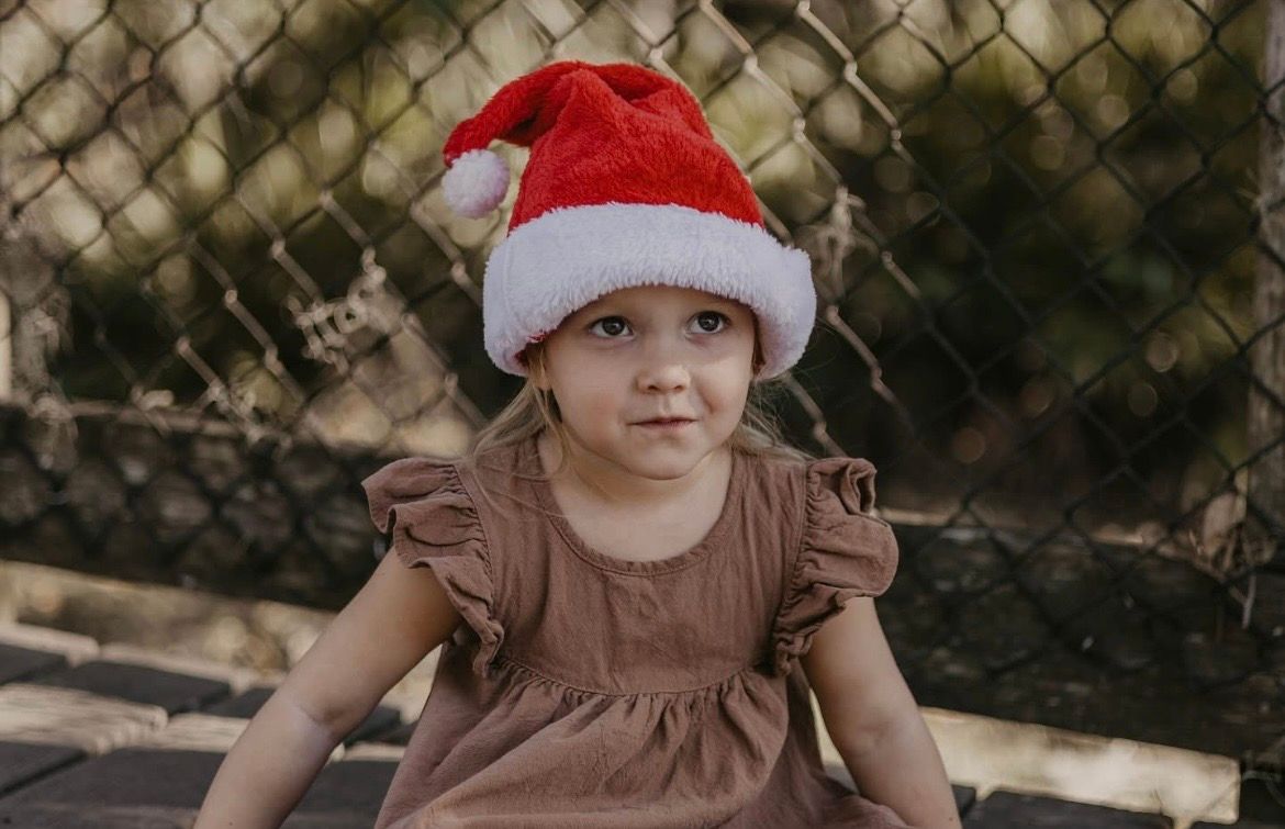 A young toddler girl wearing a red Santa hat and brown ruffle dress sits outdoors near a chain-link fence.