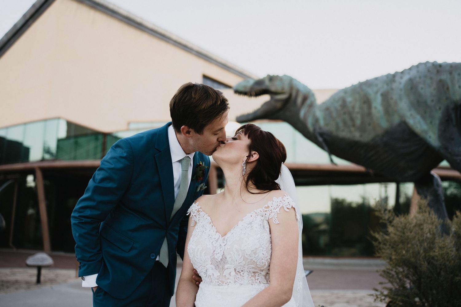 Bride and groom kissing in front of a T-Rex dinosaur statue at their unique outdoor wedding venue.