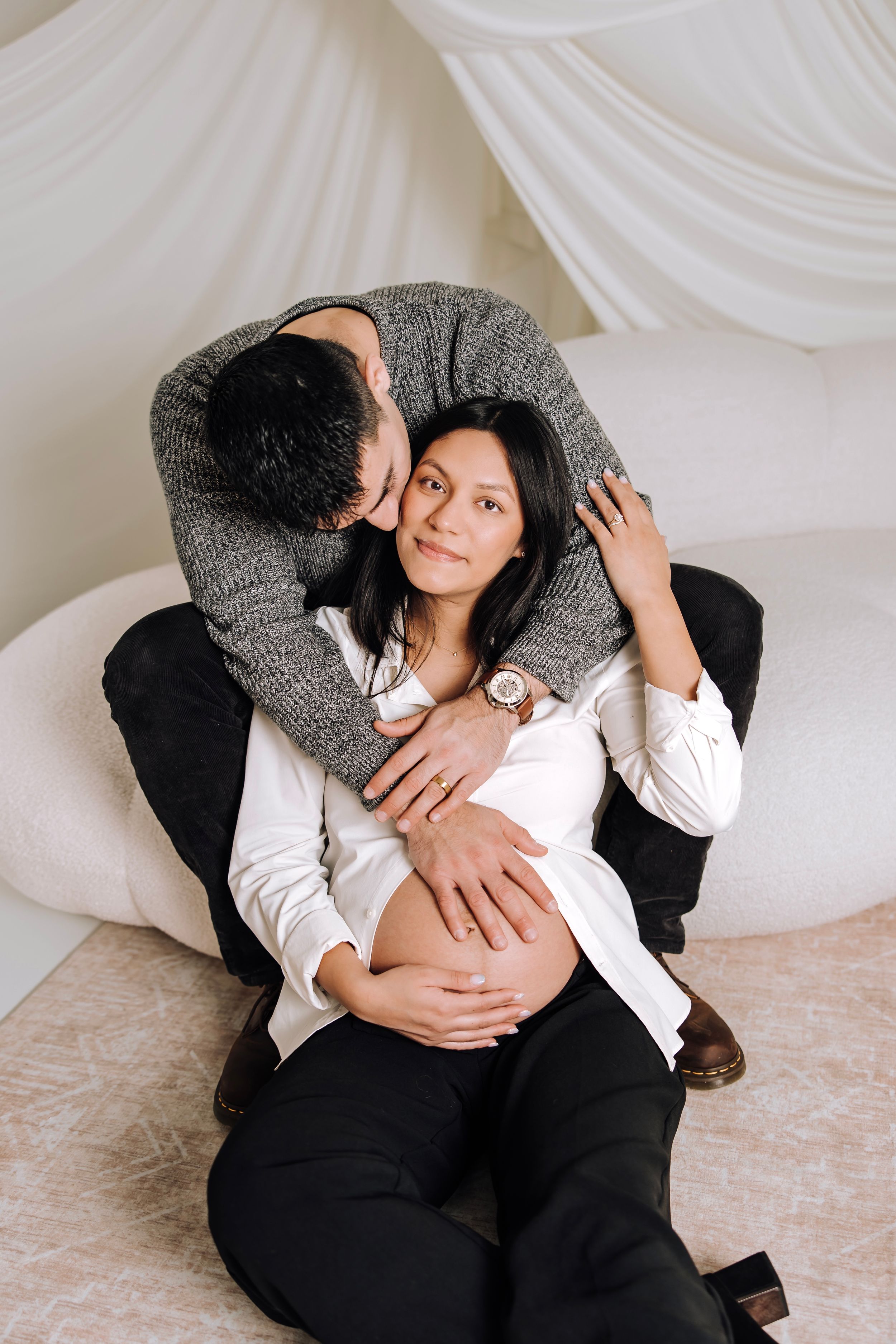 A man tenderly kisses a pregnant woman's cheek while cradling her baby bump in a softly lit white room.
