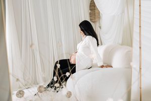 Pregnant woman in white shirt sitting on a canopy bed with white drapes, cradling her baby bump in a serene setting.