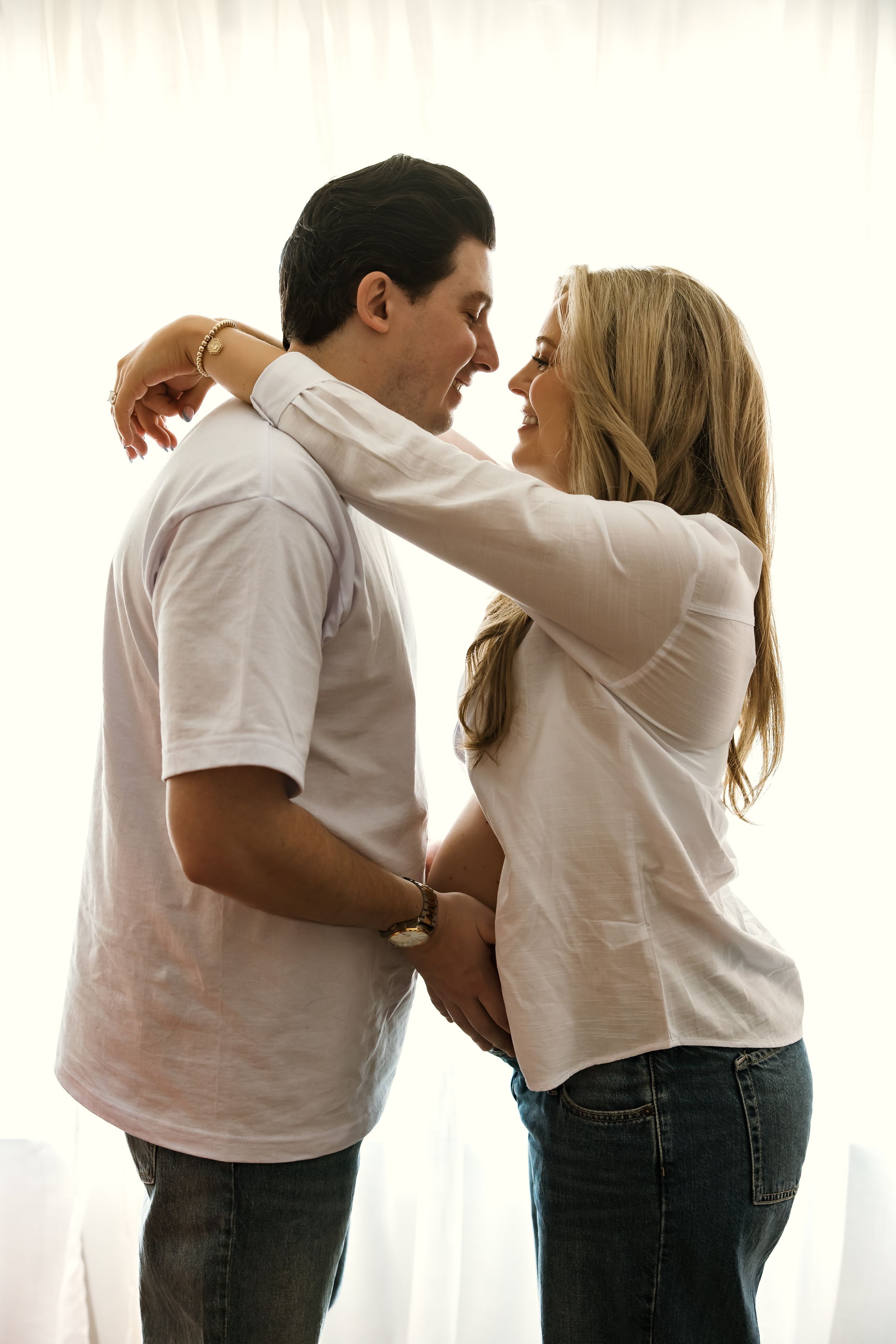 A loving couple in white shirts and jeans embrace tenderly, the woman resting her arms on the man's shoulders.
