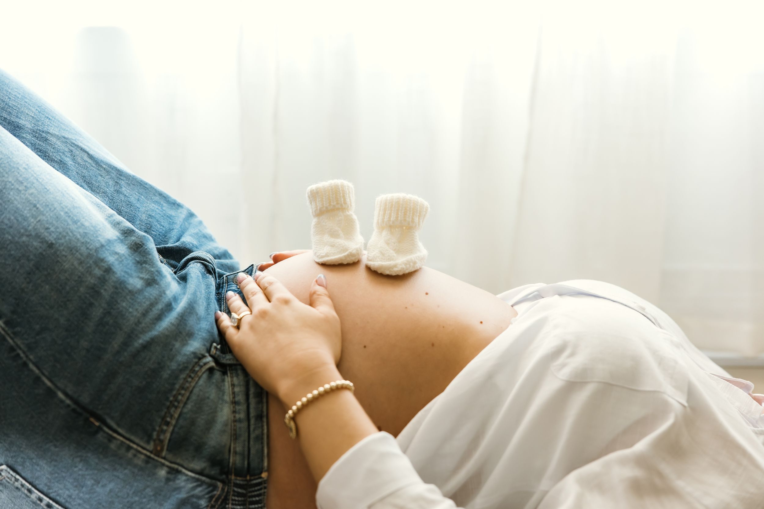 Pregnant woman lying down with tiny white baby booties resting on her bare belly, partner in jeans beside her.
