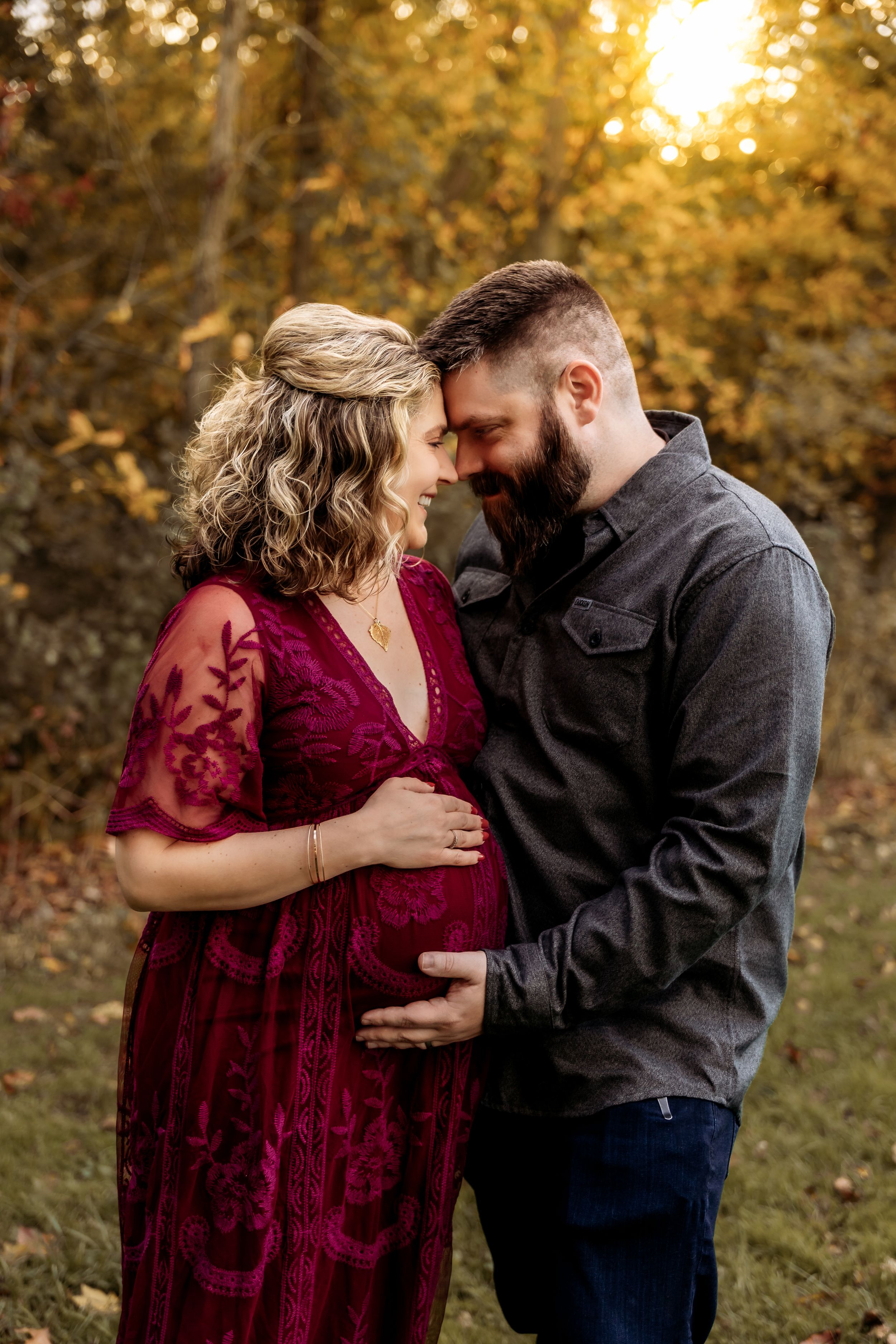 Couple sharing a tender moment outdoors during golden hour, woman in red lace maternity gown cradling baby bump.