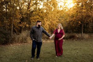 Pregnant couple holding hands in a golden autumn field at sunset, woman in red maternity gown, man in gray jacket.