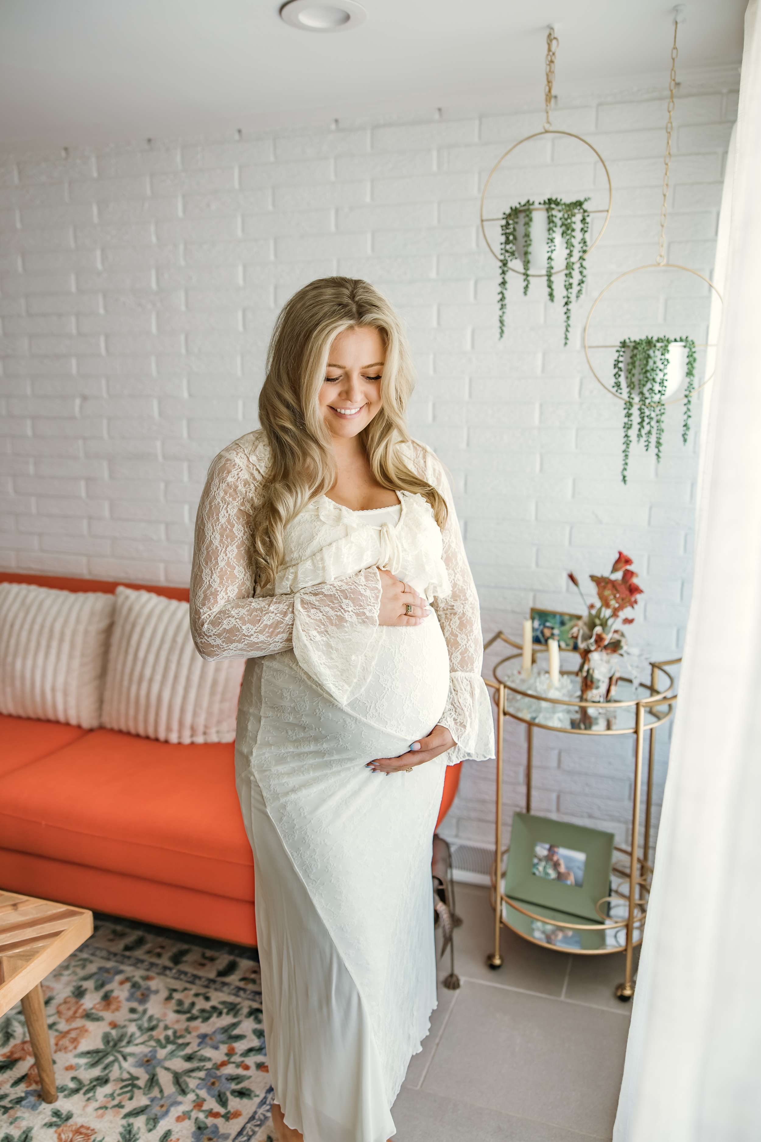 Smiling pregnant woman in white lace dress cradling her baby bump in a bright, modern living room.