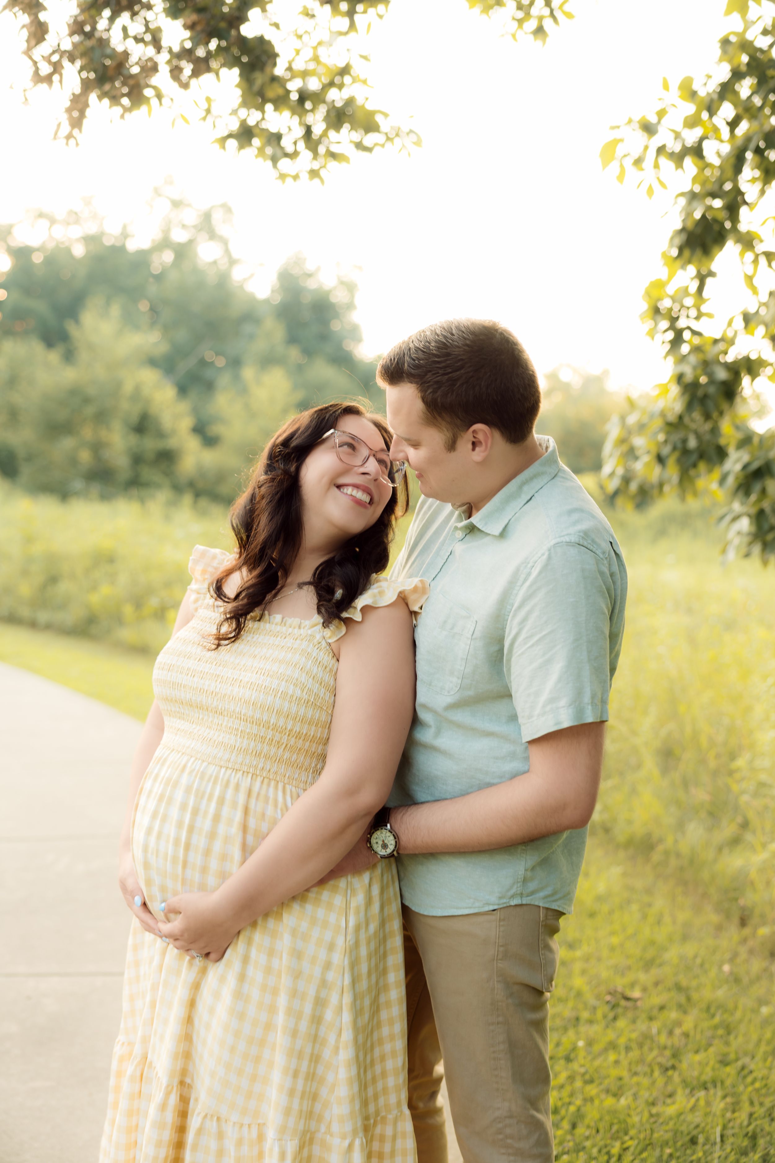 Pregnant couple smiling together outdoors in golden sunlight, woman in yellow dress cradling baby bump, man in mint shirt embracing her.