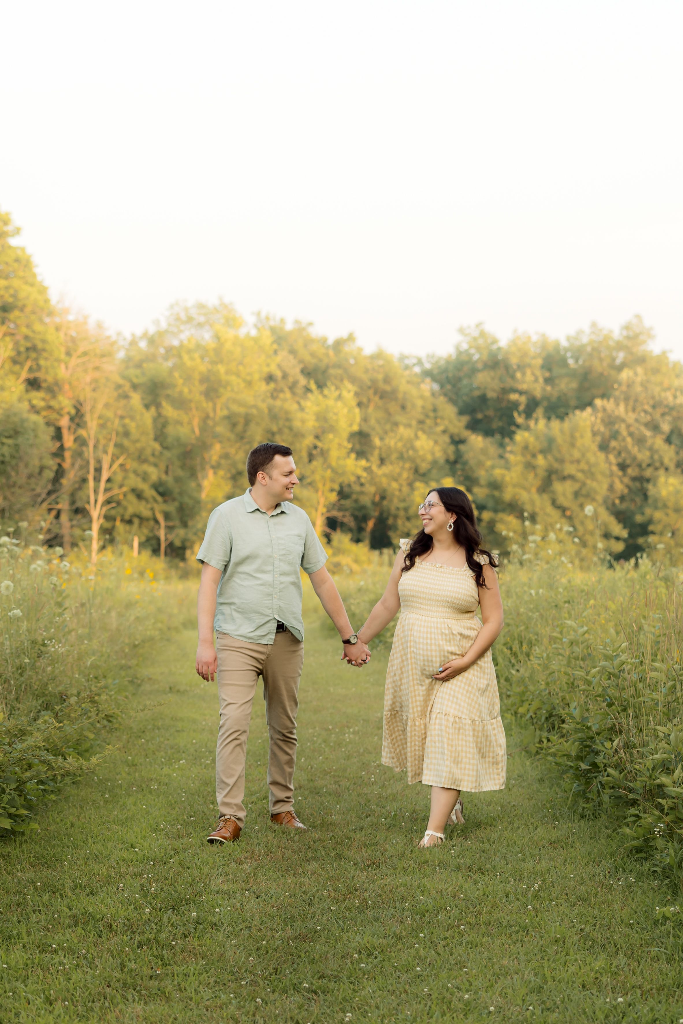 Pregnant couple holding hands and walking through a lush green meadow path during golden hour.