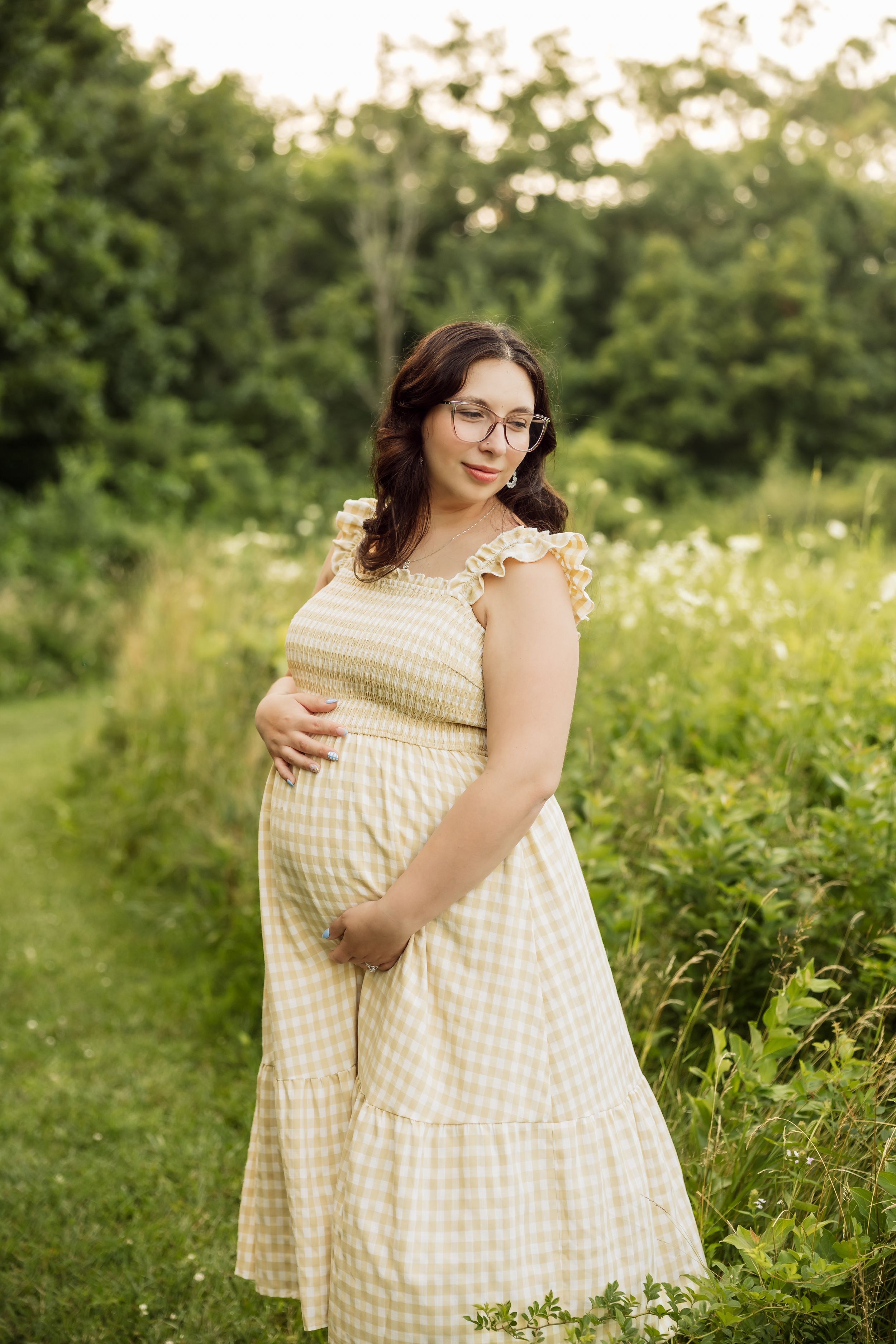 Pregnant woman in yellow gingham dress holding baby bump outdoors in lush green field during golden hour.