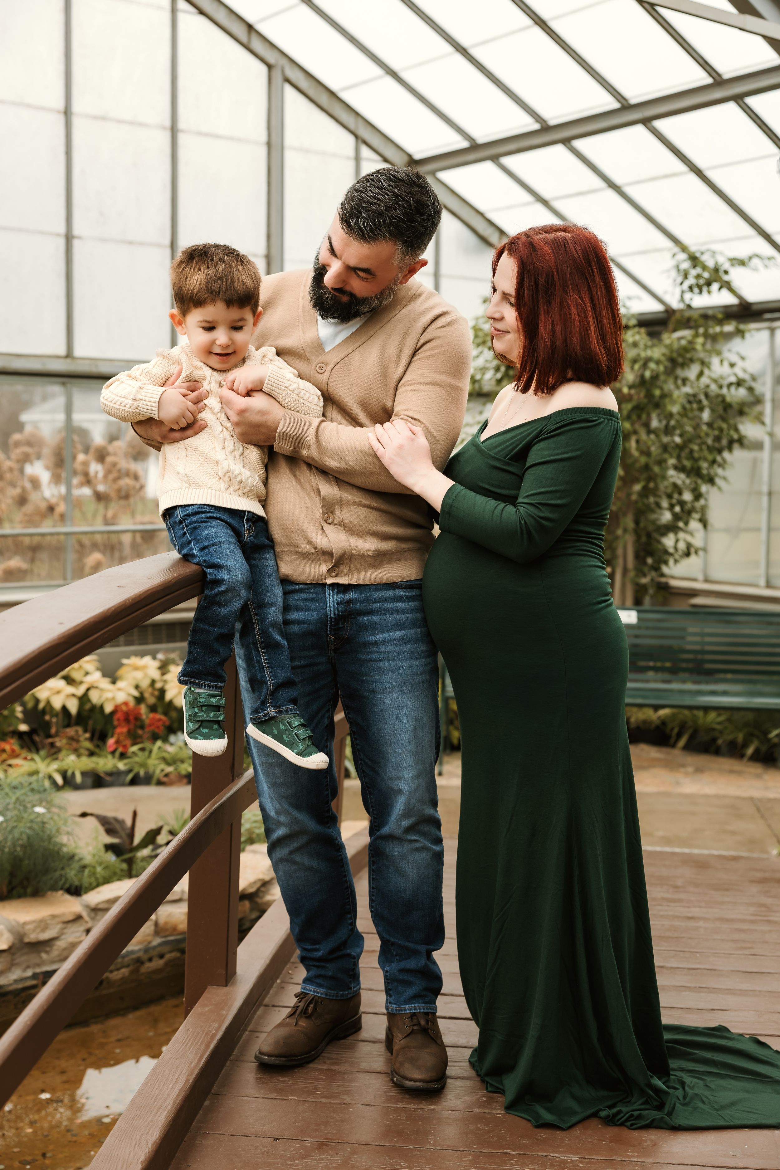 Pregnant woman in green dress with bearded man holding toddler son in a lush greenhouse setting.