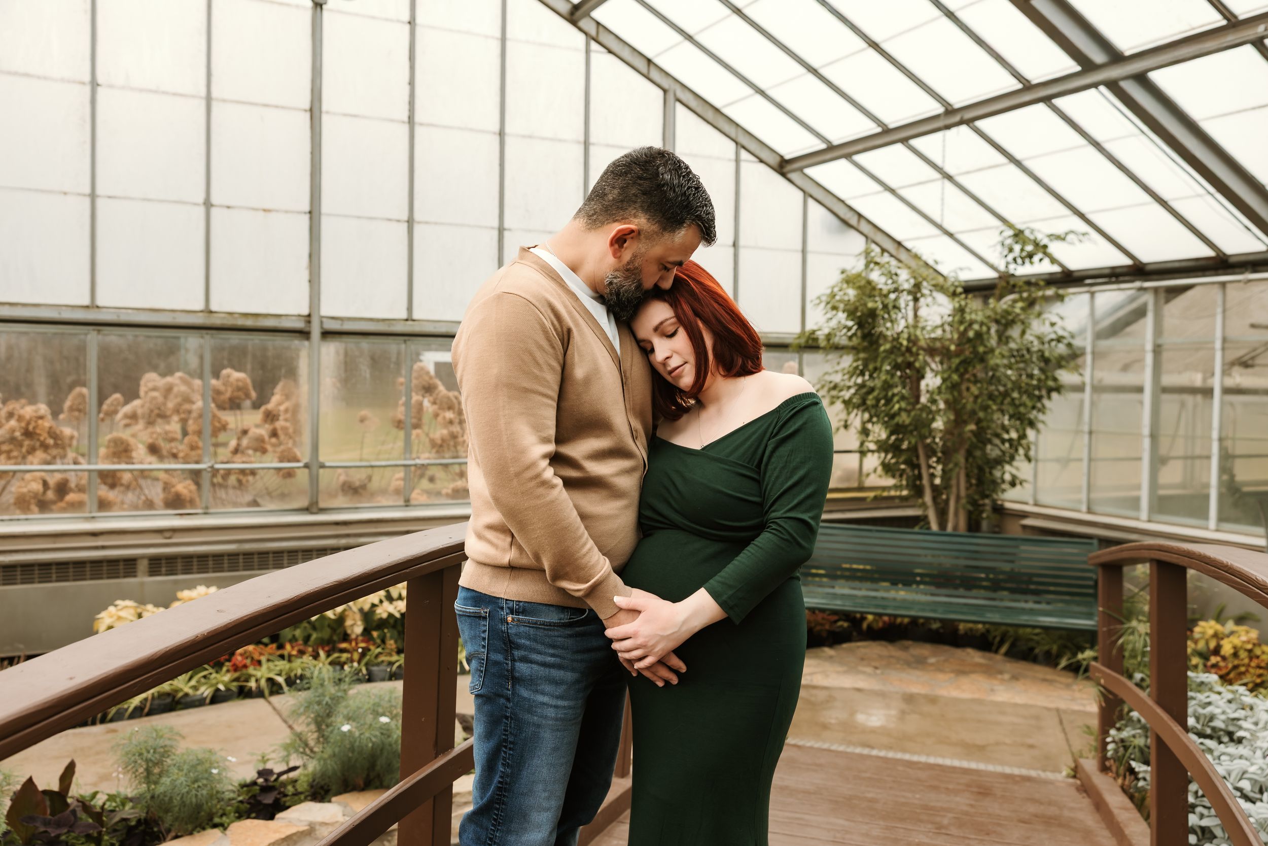Couple sharing a tender embrace in a greenhouse, woman in green dress with red hair, man in beige sweater and jeans.