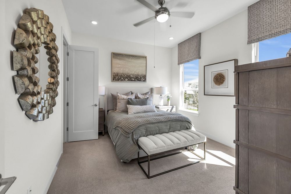 Modern bedroom with gray carpet, ceiling fan, natural light, decorative wall art, and elegant neutral-toned bedding and furnishings.