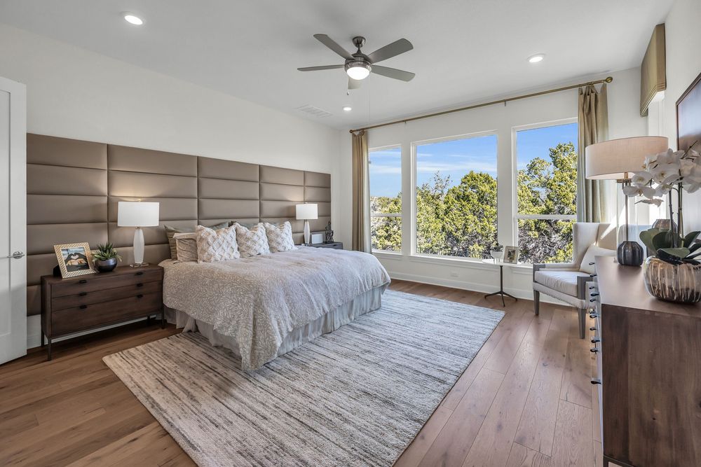 Spacious master bedroom with tall upholstered headboard, hardwood floors, large windows, and modern ceiling fan.