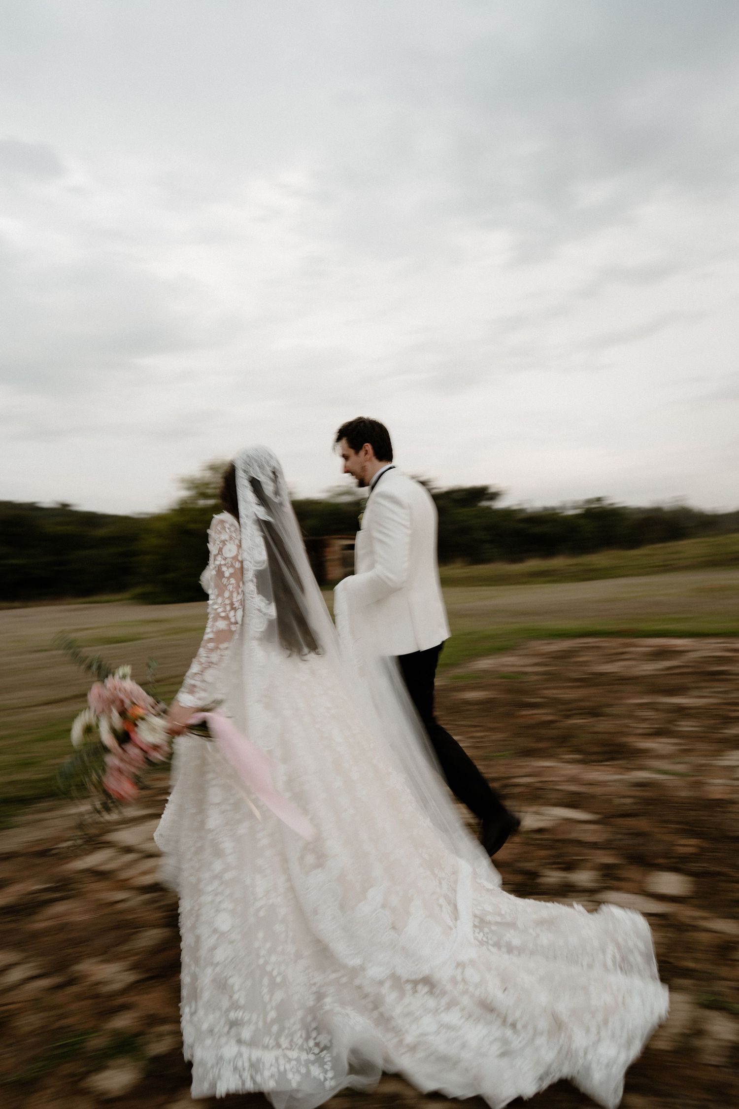 A bride and groom walk together outdoors, her lace gown and veil flowing in the wind under a cloudy sky.