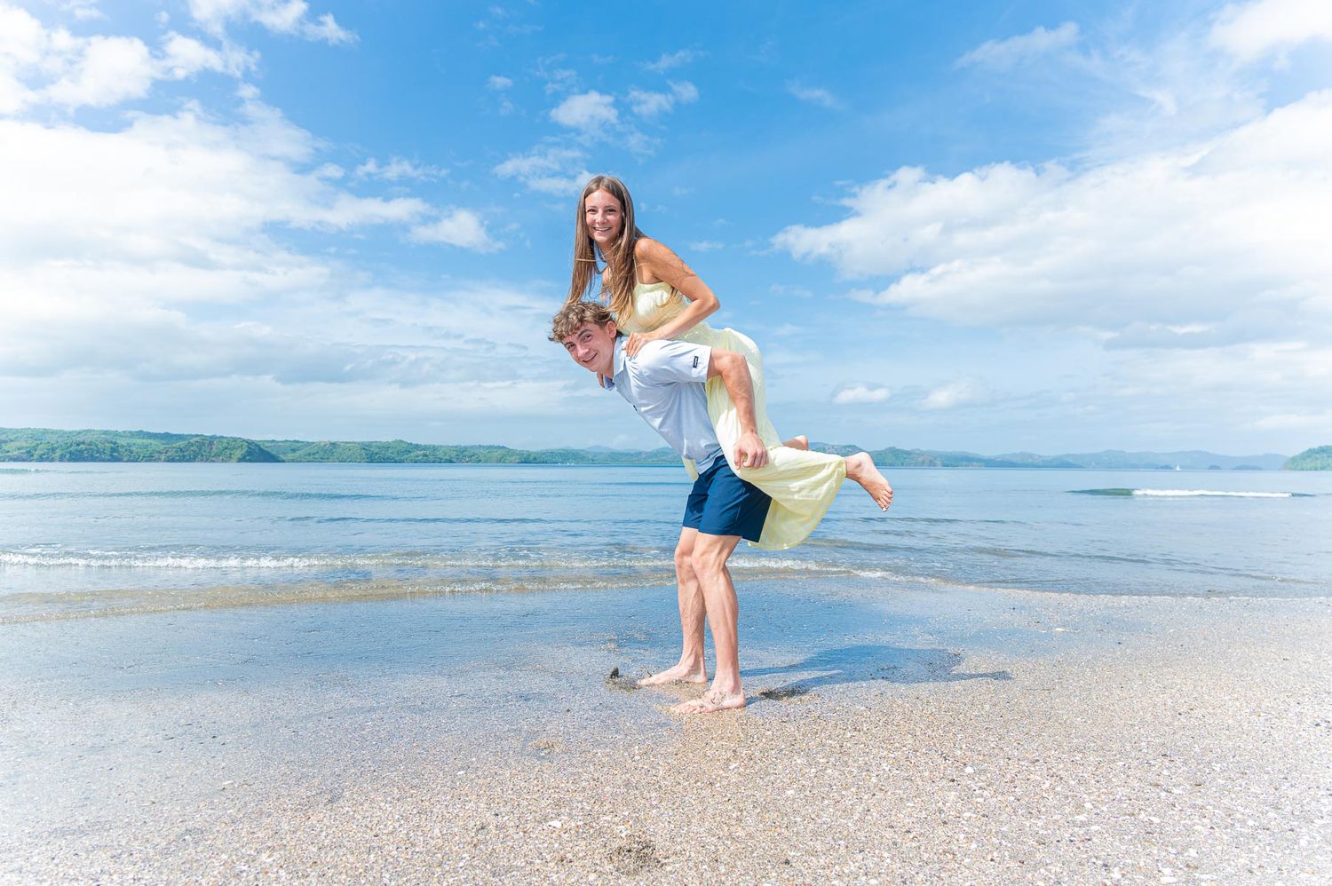 Couple playing on a sandy beach, man lifting woman in his arms with ocean waves and blue sky behind.
