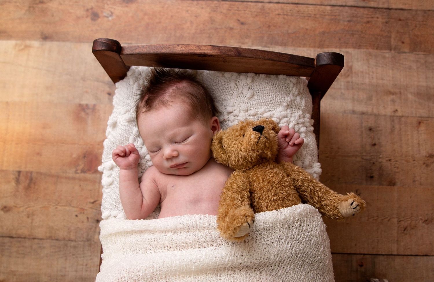 Newborn baby sleeping peacefully in a tiny wooden bed with a brown teddy bear on a rustic wood floor.
