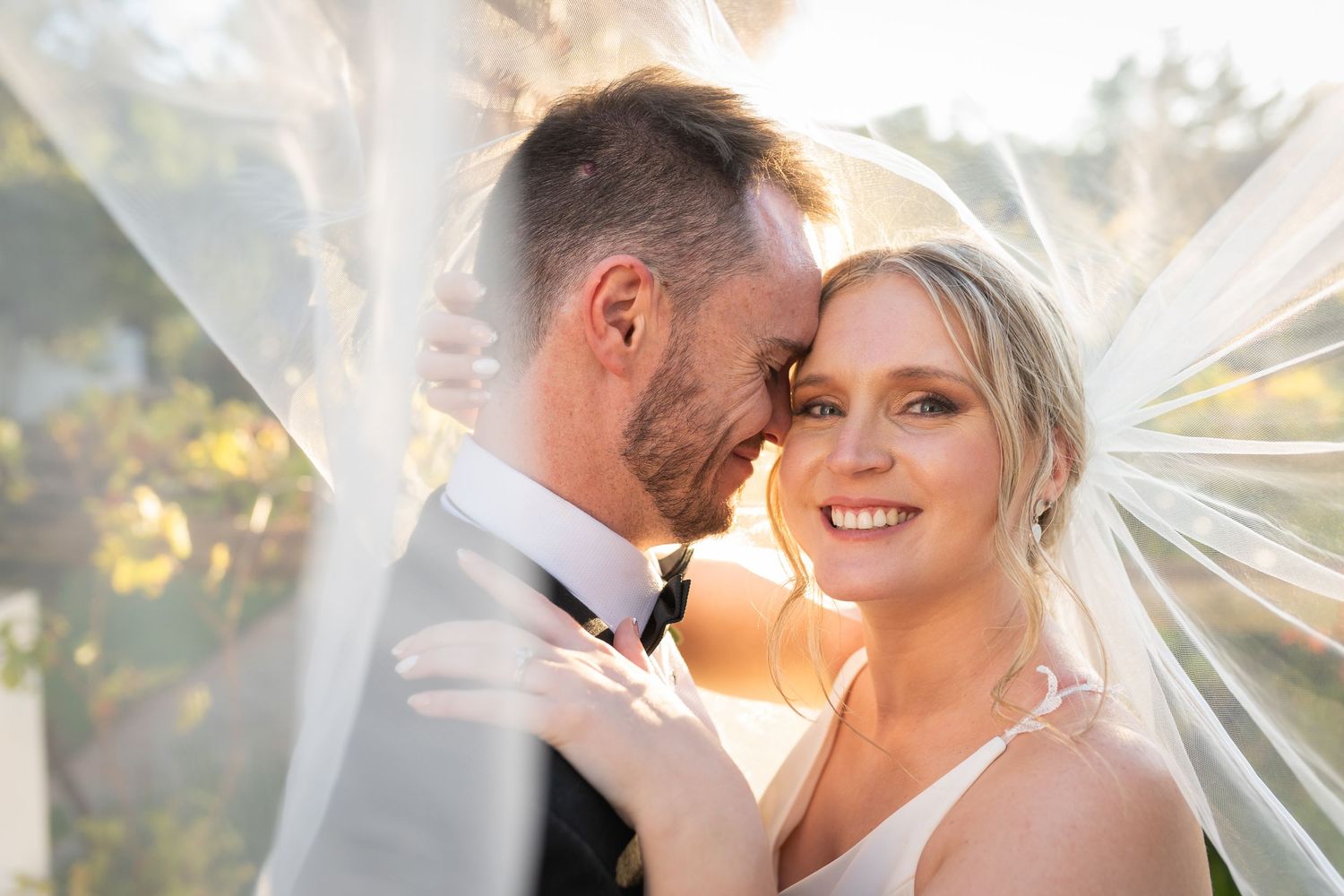 Bride and groom share a romantic moment under a flowing veil with warm golden sunlight in the background.