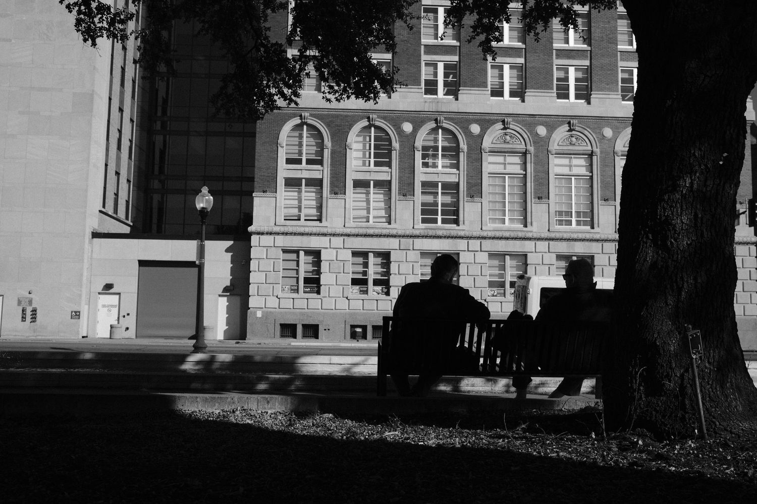 Two people sit on a bench under a tree in black and white, facing a historic brick building.