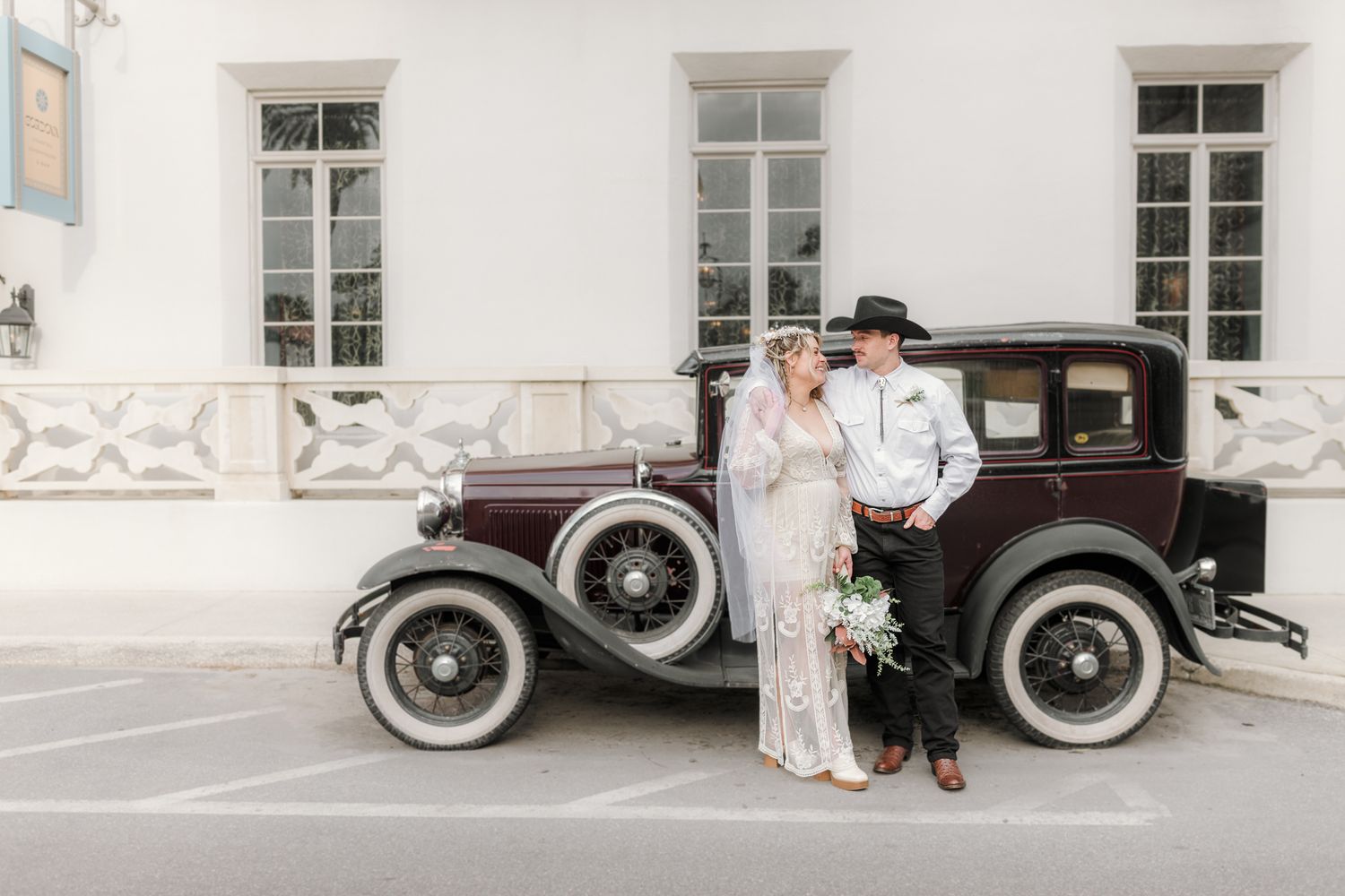 Bride and groom posing beside a vintage antique car in front of a white building on their wedding day.