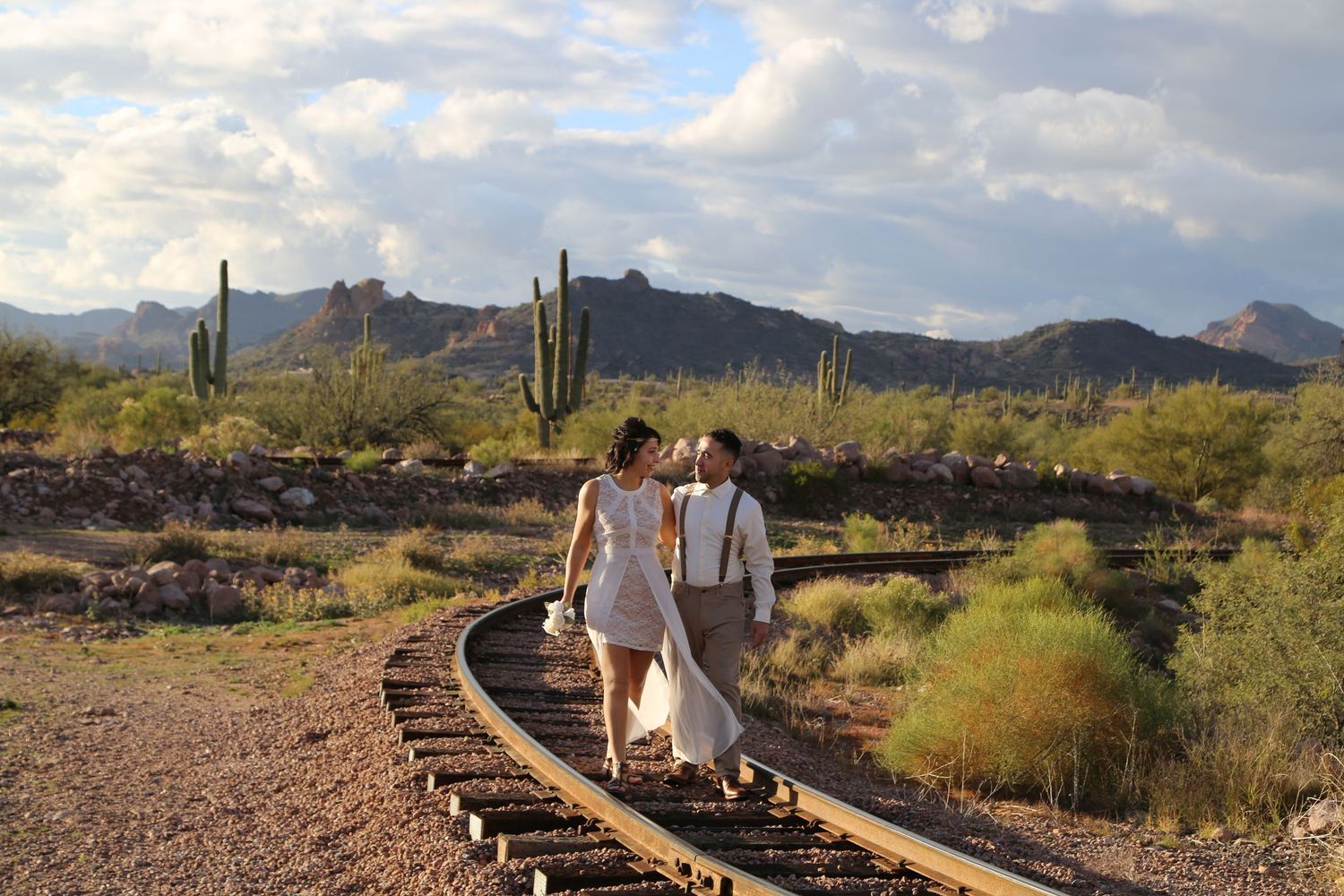 Bride and groom walk hand in hand along old railroad tracks in a desert landscape with saguaro cacti and mountains behind.