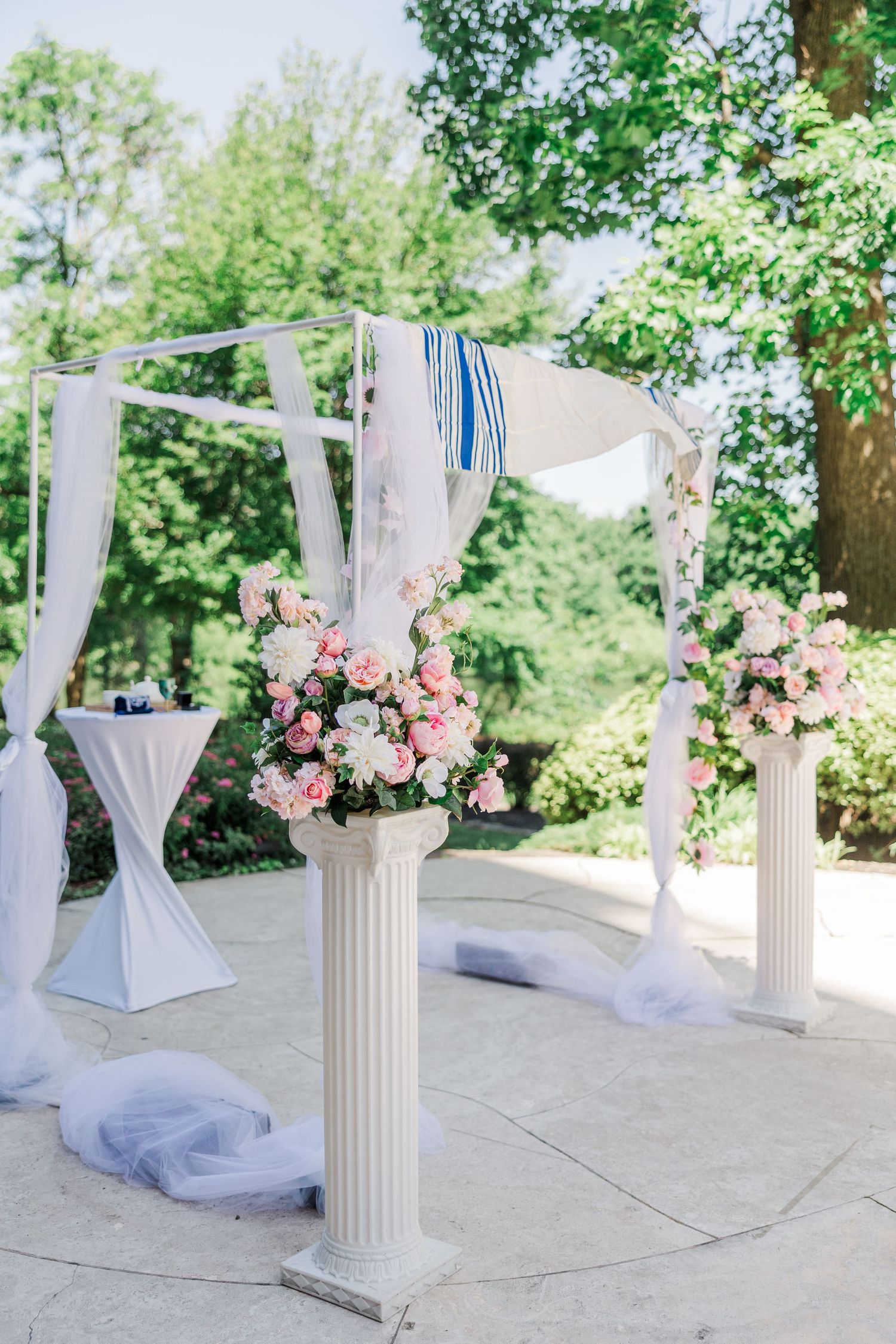 Elegant outdoor wedding ceremony arch with white draping, blue accents, and pink floral arrangements on classic white columns.