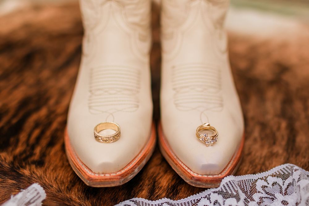 White cowboy boots with gold wedding rings displayed on top, resting on a fur surface for a rustic bridal detail shot.