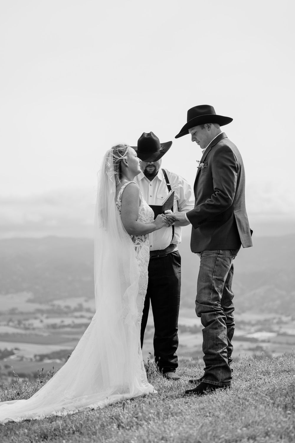 Black and white photo of a couple exchanging vows outdoors on a hilltop, officiant in cowboy hat, bride in veil, scenic valley backdrop.