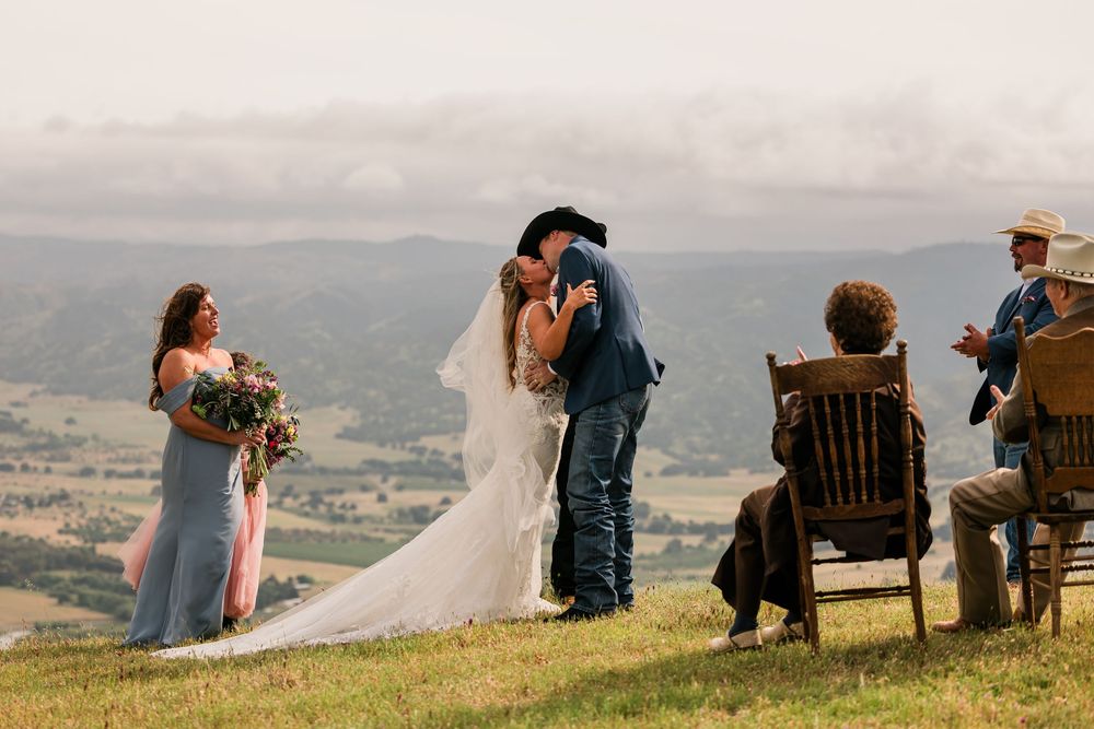 Bride and groom share first kiss at outdoor hilltop wedding ceremony with scenic mountain valley views.