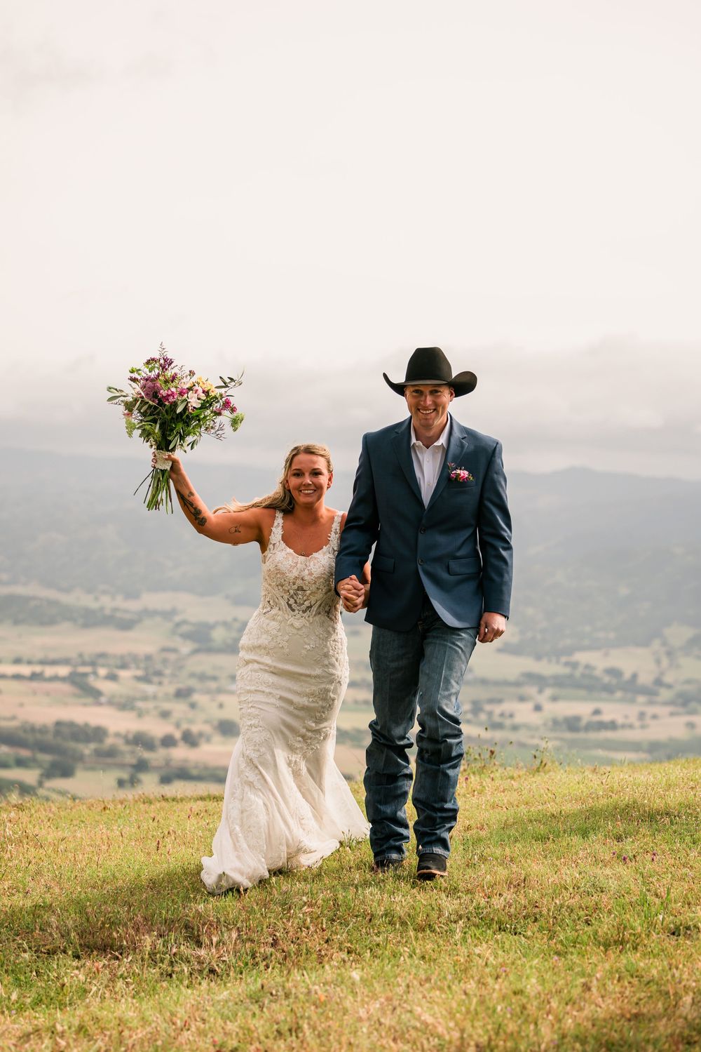 Bride in white gown holding bouquet aloft walks hand-in-hand with groom in cowboy hat and blue blazer on scenic hilltop.