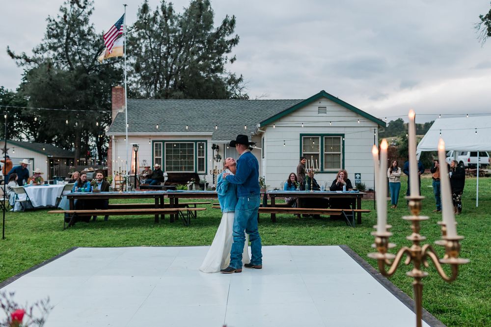 Couple shares first dance on white floor at outdoor wedding reception with American flag and white house backdrop.
