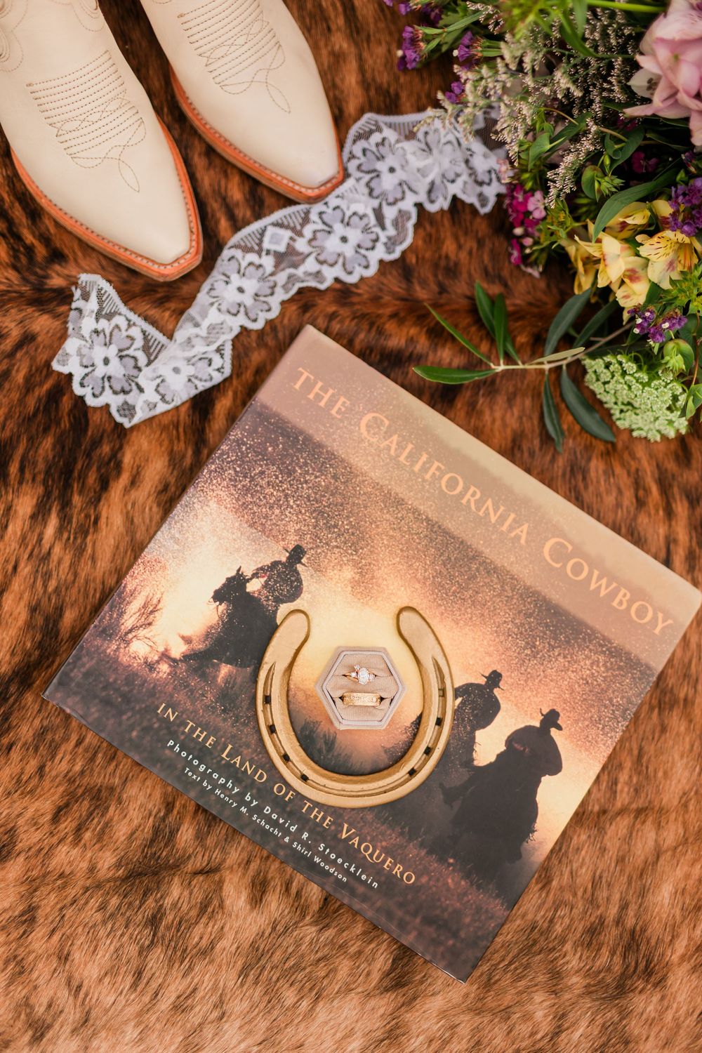 Wedding flat lay with cowboy boots, lace garter, floral bouquet, horseshoe ring dish, and The California Cowboy book.