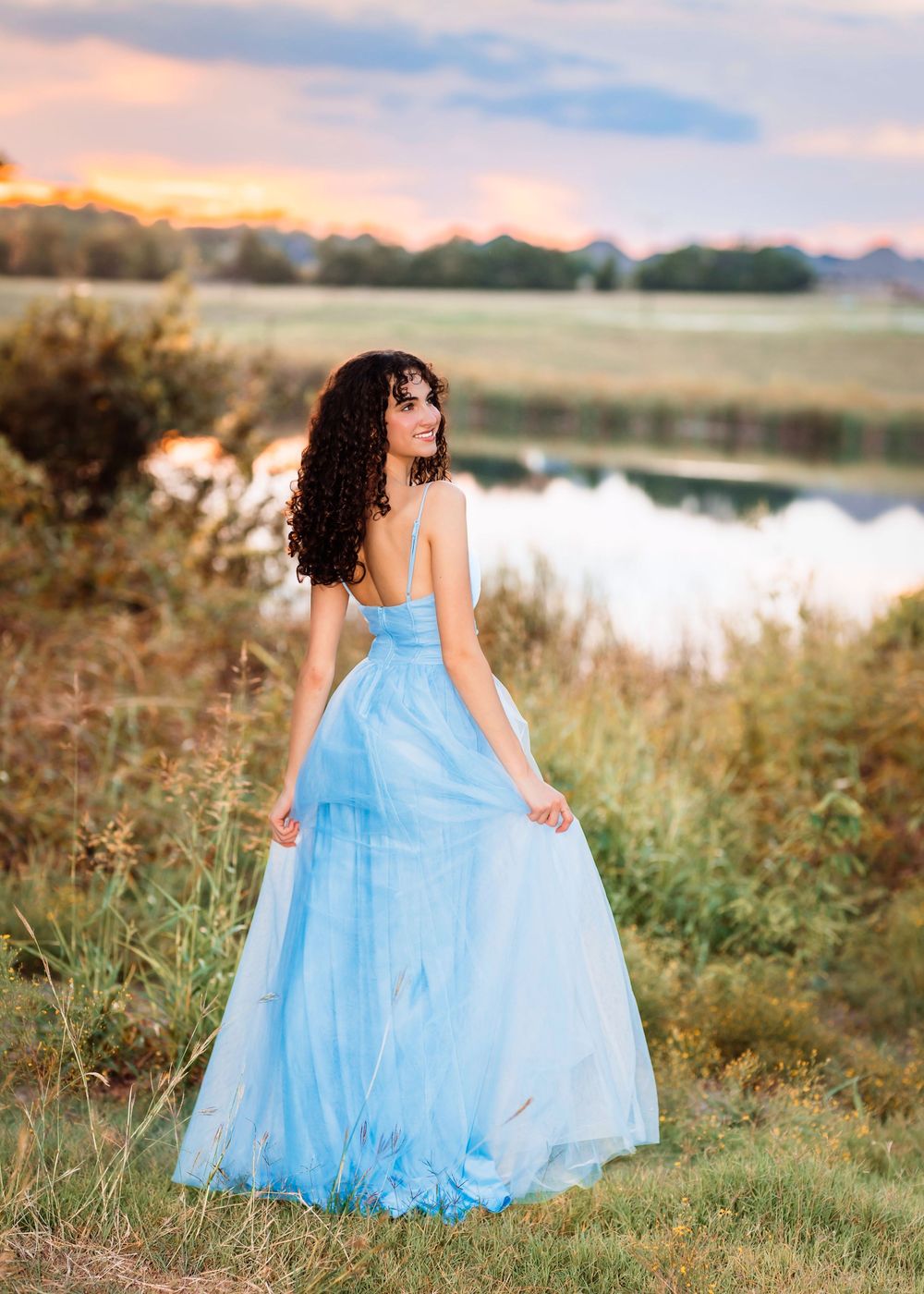 A young woman in a flowing light blue tulle gown poses outdoors near a pond at sunset with curly dark hair.