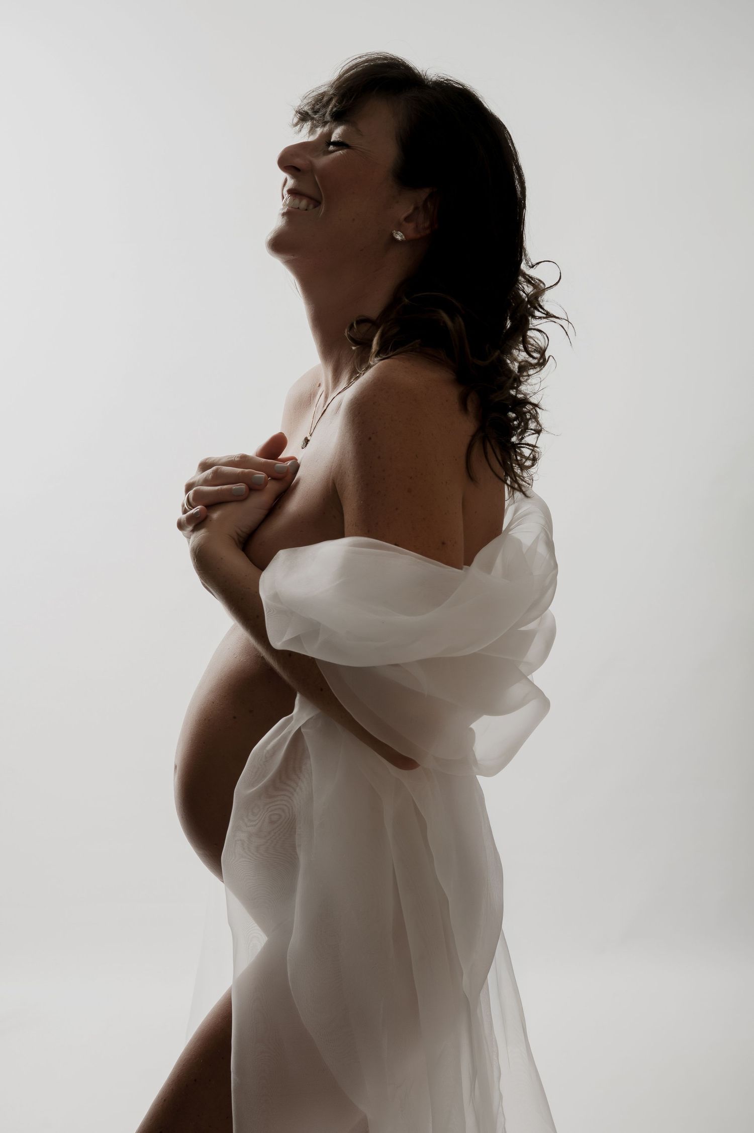 Pregnant woman draped in white fabric smiling joyfully, elegant maternity photography on light background.