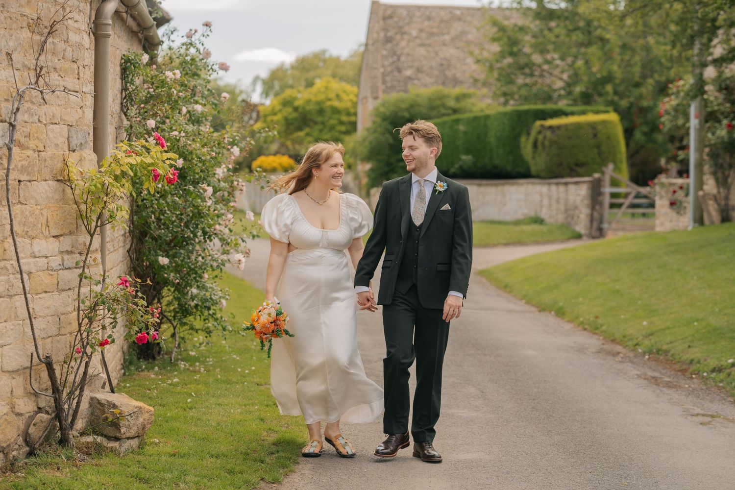 A couple stands at the entrance of a grand historic English manor house with ornate stone facade and manicured gardens.