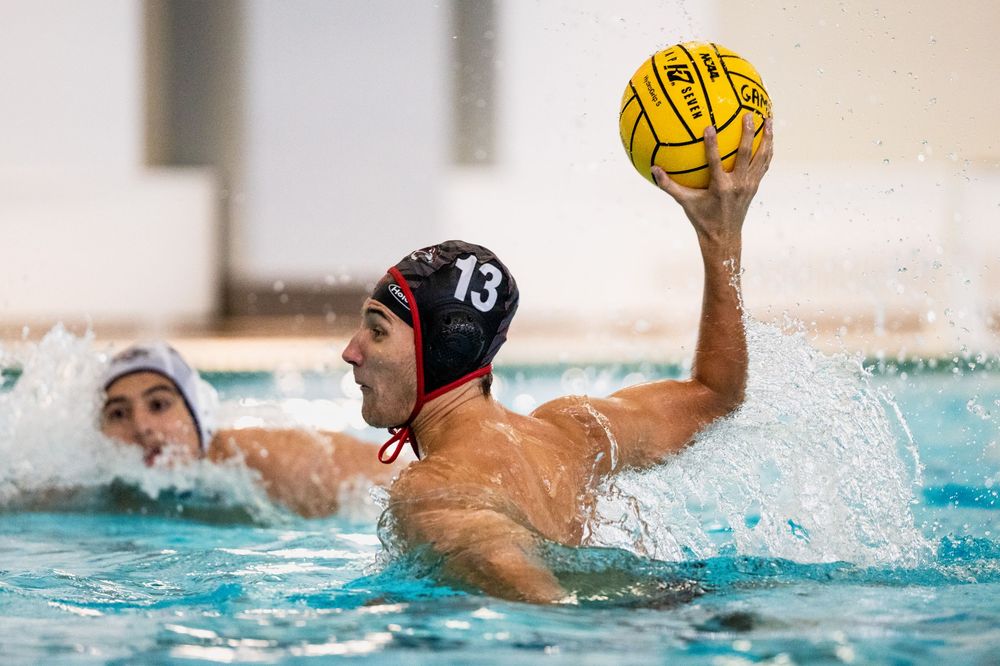 Male water polo player #13 in a red cap prepares to throw the yellow ball during a competitive match.