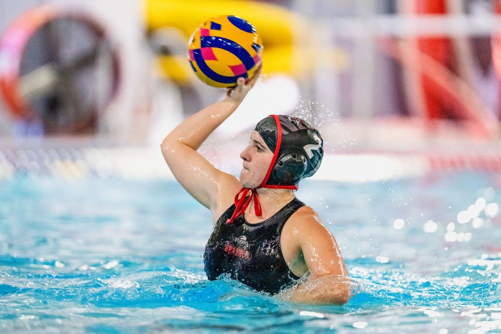 Female water polo player in a black cap prepares to throw a colorful ball during an indoor match.