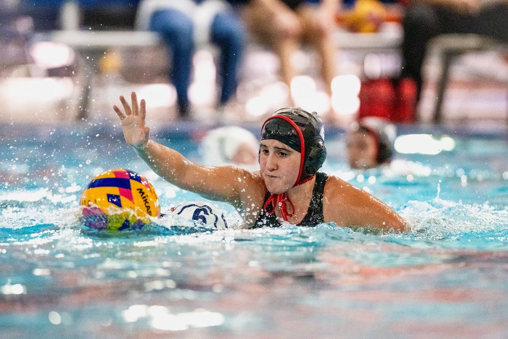 Female water polo player in a black cap reaches to block during an intense indoor pool competition.
