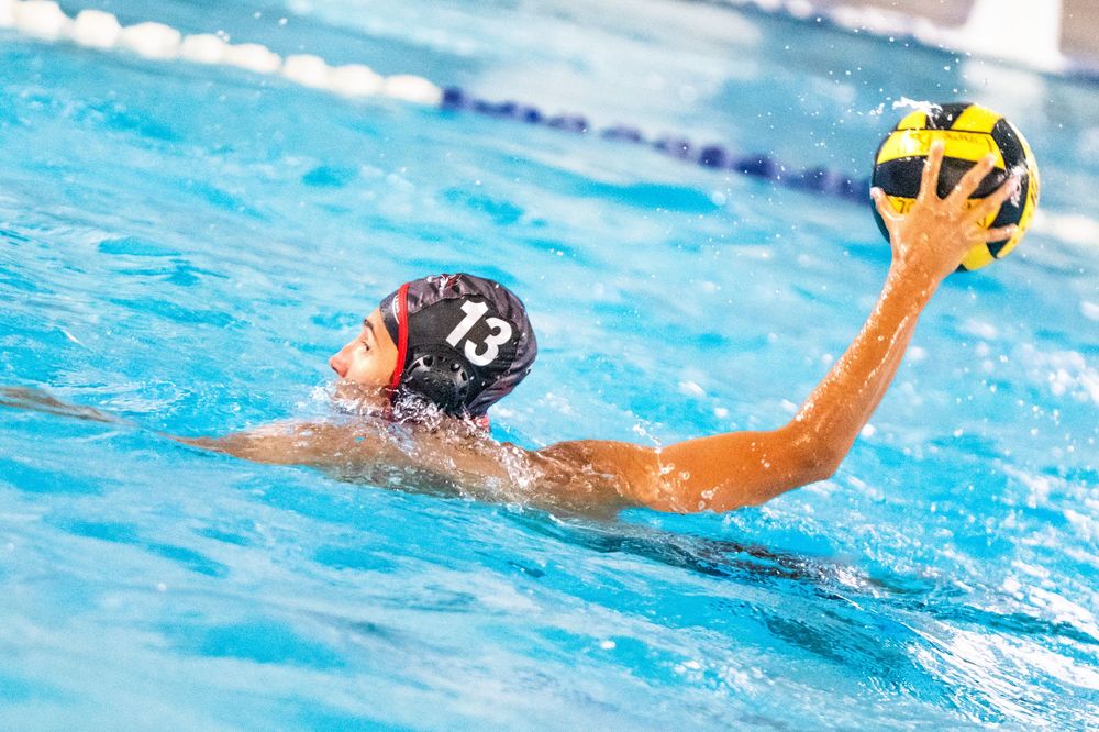 Water polo player #13 in a dark cap prepares to throw a yellow ball during an outdoor pool match.