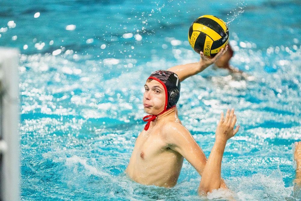 A water polo player wearing a red cap prepares to throw a yellow ball during a competitive pool match.