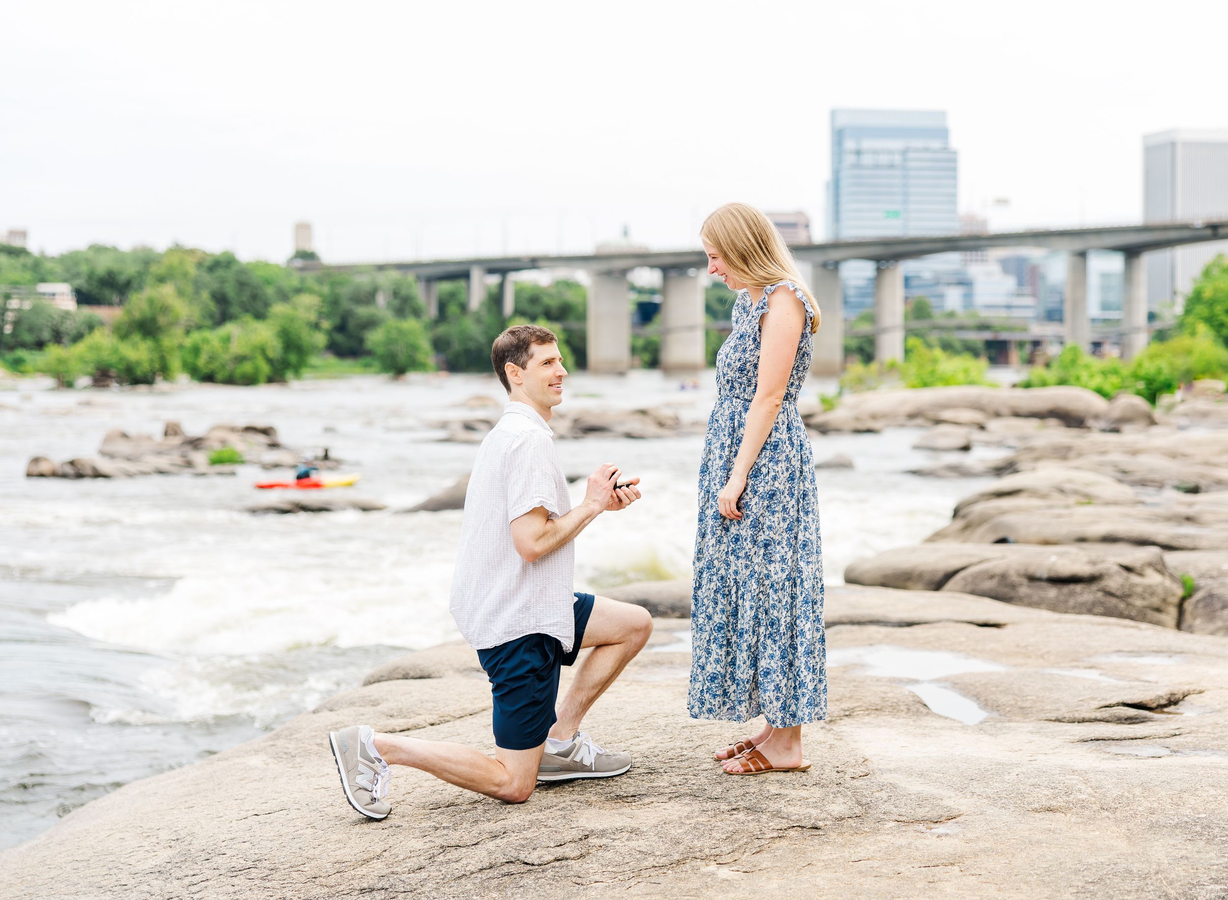 Man kneeling on rocks proposing to woman in floral dress by river with city bridge backdrop.