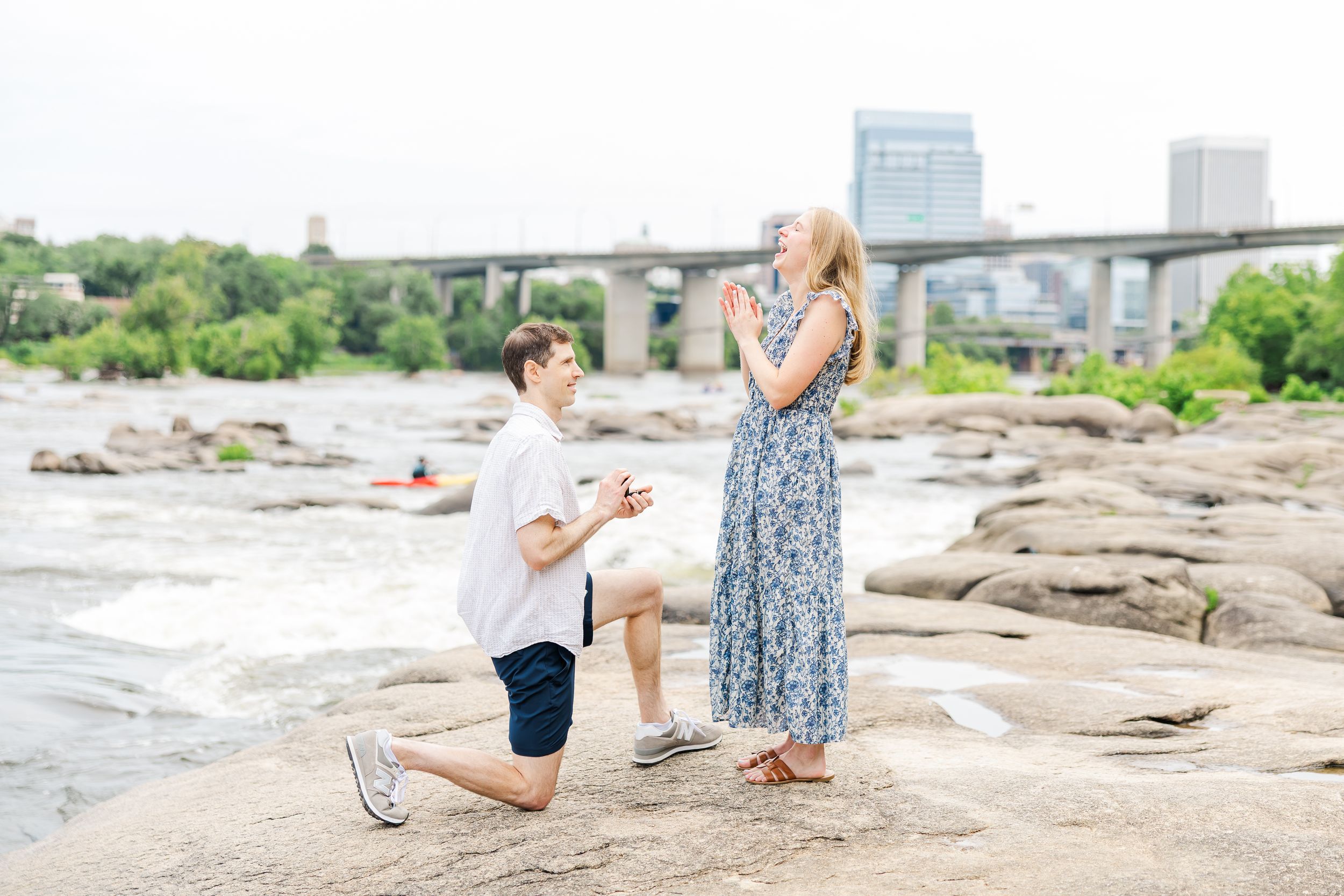 Man kneeling on rocks proposing to woman in floral dress by a river with a bridge in the background.