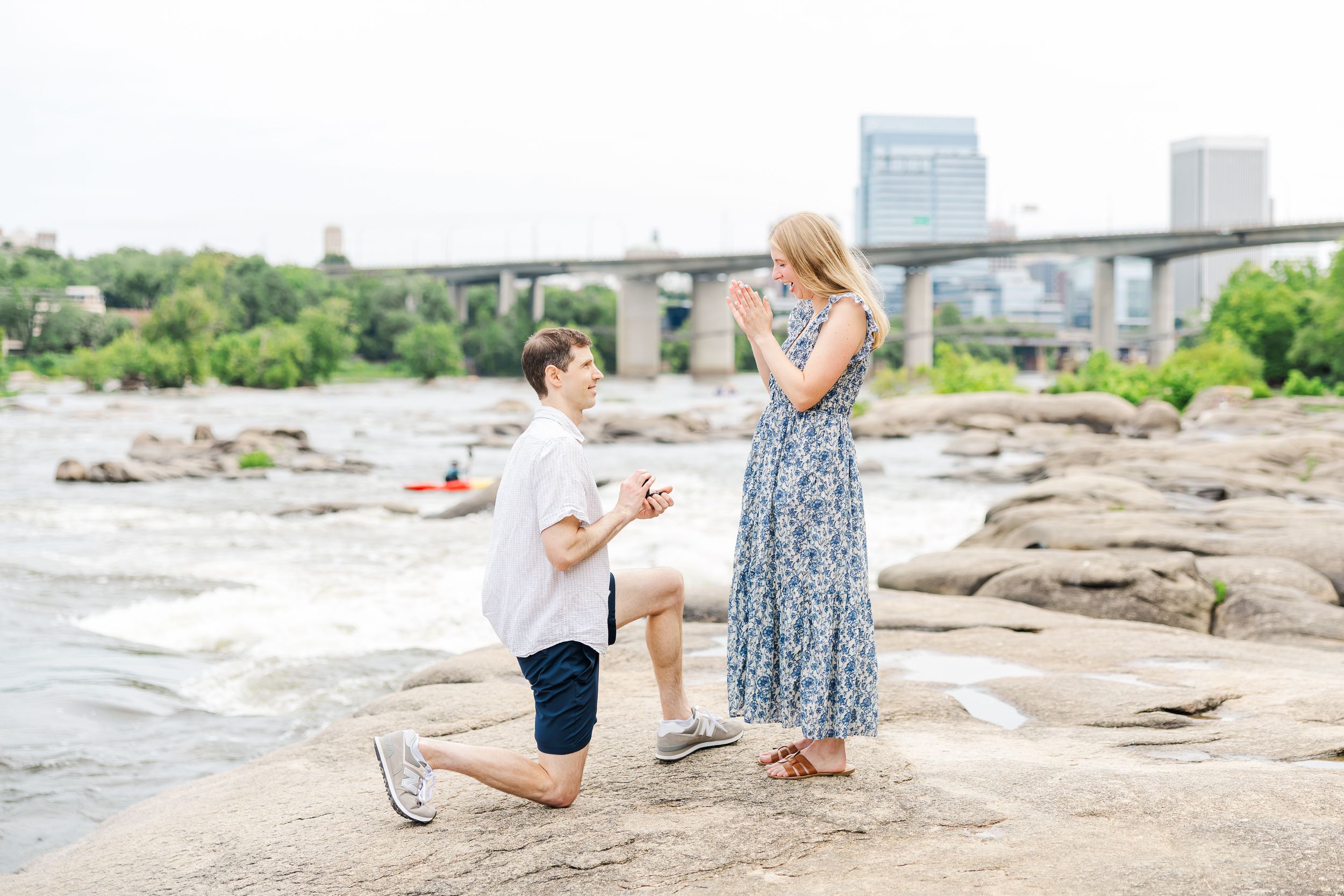 Man proposing to woman on rocky riverbank with bridge and rapids in background.